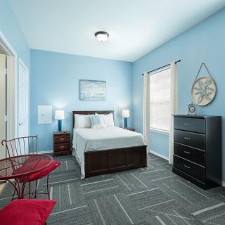 A modern bedroom with blue walls, featuring a bed, nightstands with lamps, a dark dresser, and a small table with red chairs.