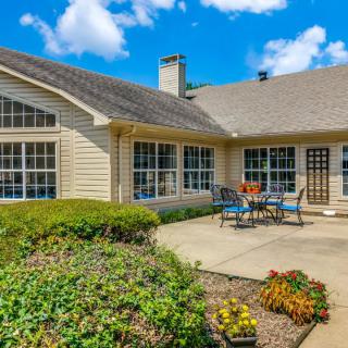 A cozy patio with outdoor furniture in front of a house, surrounded by a garden on a sunny day.