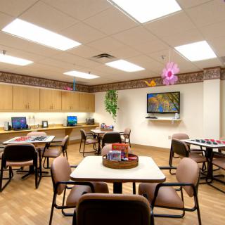 A well-lit room with multiple tables and chairs, featuring a television and computer workstations along the wall.