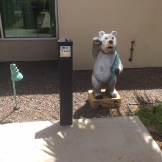 A bear statue stands on a sidewalk next to a tall black post on a sunny day.