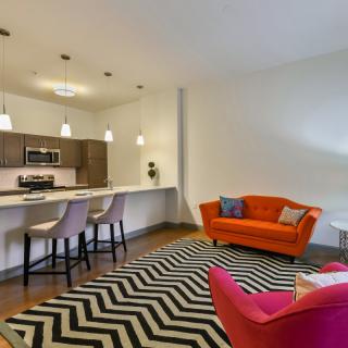 A modern open-concept living space with a vibrant orange couch and zigzag patterned rug, seamlessly connected to the kitchen area.