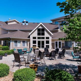 A spacious outdoor patio area with tables and chairs in front of a large building, surrounded by well-maintained landscaping under a clear blue sky.