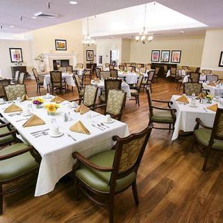 An elegantly set dining room with neatly arranged tables and chairs, featuring white tablecloths and a wooden floor.
