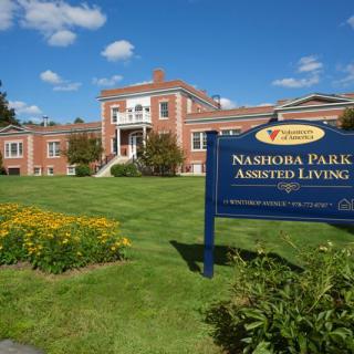 A scenic view of Nashoba Park Assisted Living with a brick building and manicured lawn.