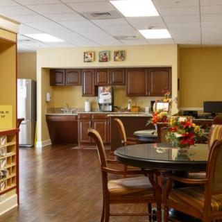 A cozy dining area with wooden furniture and a kitchen in the background, decorated with flowers on a round table.
