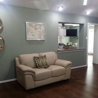 A modern living room with a beige sofa against a green wall, decorated with wall art and mirrors, featuring an open view to the kitchen area.