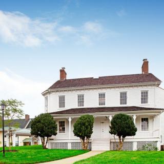 A large, two-story white house with a porch and surrounding greenery under a clear blue sky.