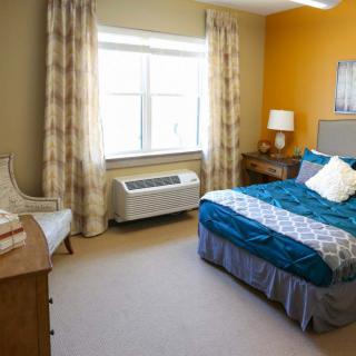 A cozy bedroom with a neatly made bed, complete with vibrant blue and white bedding, positioned next to a window adorned with patterned curtains. The room includes a chair, wooden dresser, and a bedside lamp for added comfort.