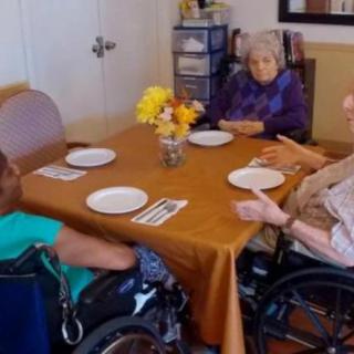 Seniors in wheelchairs sit around a dining table, engaged in conversation.