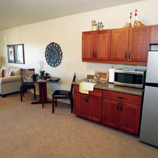 A cozy living space with a kitchenette, including wooden cabinets, a microwave, and a refrigerator, adjoined to a sitting area with a sofa and small dining table.