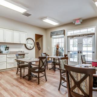 A cozy kitchen and dining area with wooden floors, featuring several tables and chairs, a clock on the wall, and bright natural lighting.