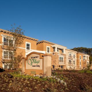 A sunny view of a residential apartment building with a landscaped entrance and prominent signage.