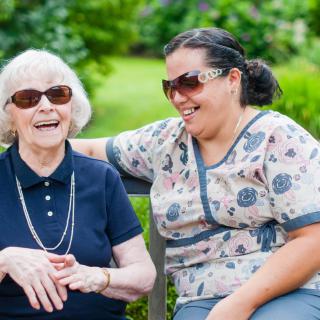 An elderly woman and her caregiver share a joyful moment outdoors, both wearing sunglasses and smiling.