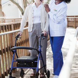 A caregiver assists an elderly person using a walker outdoors on a wooden ramp.
