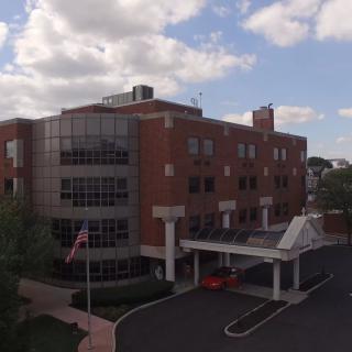 A brick hospital building with a covered entrance and parking area under a cloudy sky.