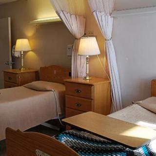 A hospital room with two beds separated by a curtain, featuring wooden furniture and lamps.