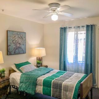 A cozy bedroom with a single bed adorned with a green and gray quilt, featuring a lamp, curtains, and a decorative painting.
