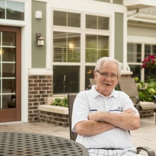 An elderly man sitting outside on a patio in front of a residential home.