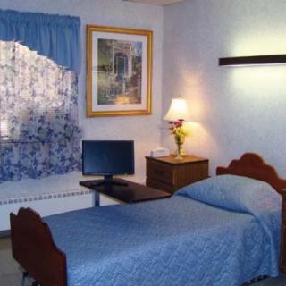 A small bedroom featuring a single bed with blue bedding, a lamp on a nightstand, and a computer monitor.
