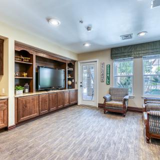 A cozy living room with wooden flooring, a television, bookshelves, and comfortable seating near large windows.