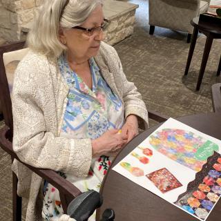An elderly woman wearing glasses is seated at a table, observing some artwork.