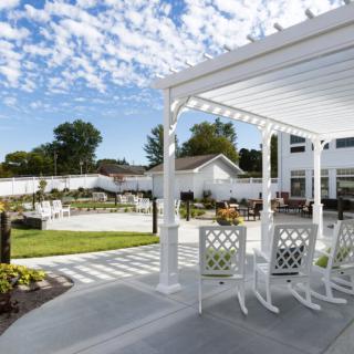 A serene backyard patio with a pergola, featuring white rocking chairs and a well-maintained lawn under a bright, sunny sky.
