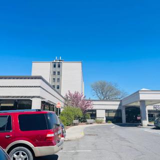 A view of a hotel building with trees and vehicles parked in the lot under a clear blue sky.