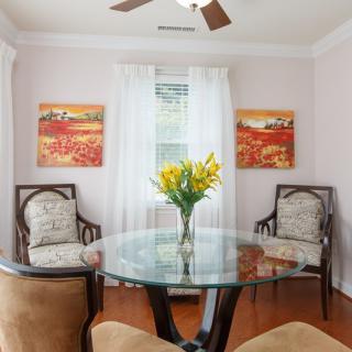 A cozy living room features a glass table surrounded by elegant chairs, with a vibrant floral arrangement and colorful artwork adding charm.