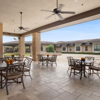 An outdoor patio with tables and chairs, covered by a roof with ceiling fans, offering a view of a landscaped area.