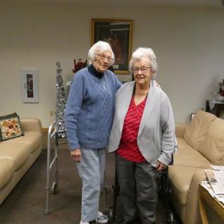 Two elderly women standing together in a living room, showcasing friendship and warmth.