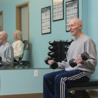 An elderly man exercises in a gym, lifting dumbbells while seated and looking at the mirror.