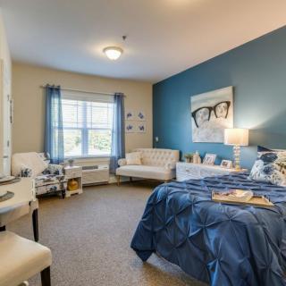 A cozy bedroom with a blue accent wall, featuring a bed, comfortable seating, and a window allowing natural light.