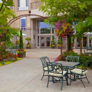 A charming outdoor patio area with a table and chairs surrounded by lush greenery and colorful flowers in front of a building.