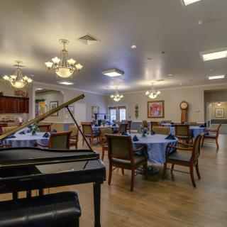 A spacious dining room featuring elegantly set tables, comfortable chairs, and a grand piano under warm chandeliers.