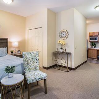 A cozy bedroom with stylish decor, featuring a bed, decorative chair, and a small side table, with a kitchenette in the background.