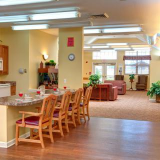 A bright kitchen and dining area with wooden chairs and tables, featuring plants and warm lighting.