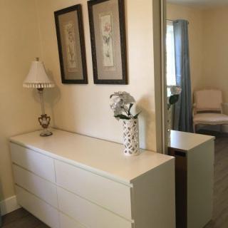 A bedroom corner featuring a white dresser topped with a decorative lamp and vase, complemented by framed art and a large mirror.