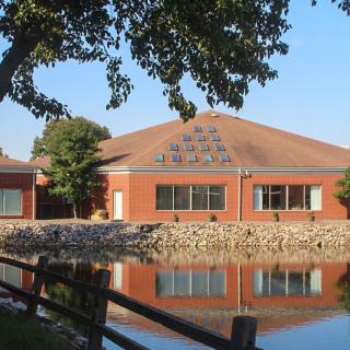 A building with a gabled roof is reflected in a calm lake, surrounded by trees.