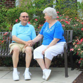 An elderly couple sits on a park bench surrounded by roses, holding hands and enjoying a conversation.