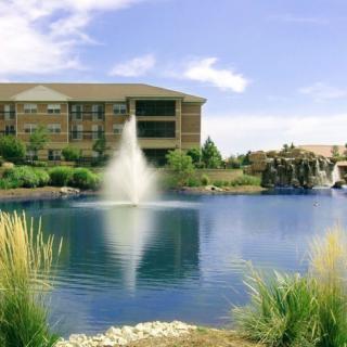A serene pond with a fountain in front of a building, surrounded by lush greenery and a clear sky.