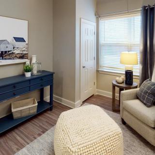A cozy living room corner with an armchair, a pouf, and a blue cabinet, illuminated by a lamp near the window.