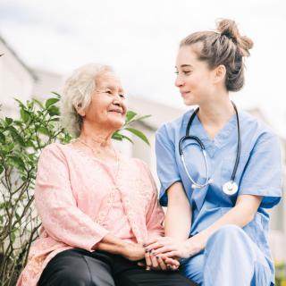 A nurse in scrubs holds hands with an elderly woman, both smiling while sitting outdoors.