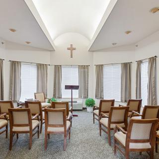 A small chapel interior with chairs arranged in rows, a podium at the front, and a cross on the wall.