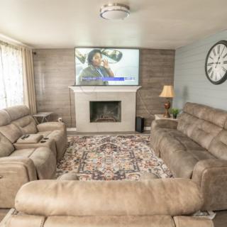 A cozy living room featuring a large sofa set, a television above the fireplace, and elegant decor elements like a patterned rug and wall clock.