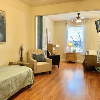 A spacious living room featuring a bed, comfortable chairs, and a large window allowing natural light to illuminate the wooden floors.