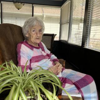 An elderly woman relaxes in her cozy indoor space, dressed in comfortable plaid pajamas.