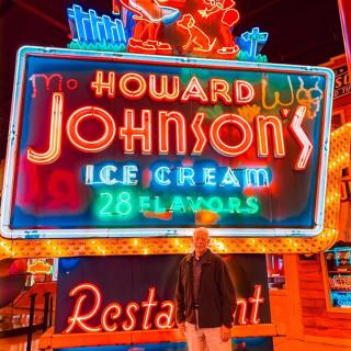 A person standing in front of a brightly lit Howard Johnson's neon sign advertising ice cream and a restaurant.