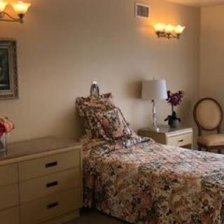 A cozy bedroom with floral bedding, featuring a single bed, nightstand, lamp, and chair.