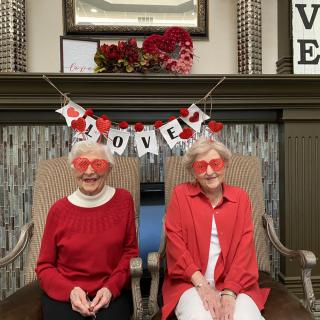 Two women wearing red outfits and heart-shaped glasses sit under a 'LOVE' banner, surrounded by festive decorations.