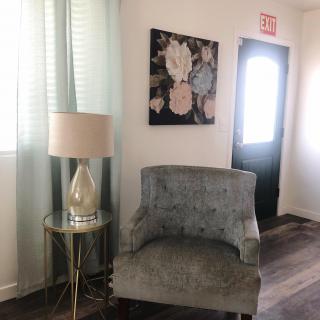 A cozy living space featuring a plush chair, a small side table with a lamp, and floral artwork on the wall.